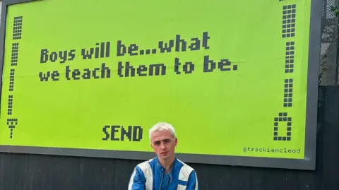 Trackie McLeod Trackie McLeod with bleach blonde hair and a blue jacket, stands in front of a large neon green billboard. The billboard is in the style of a retro Nokia phone screen, with lettering that reads "boys will be... what we teach them to be."