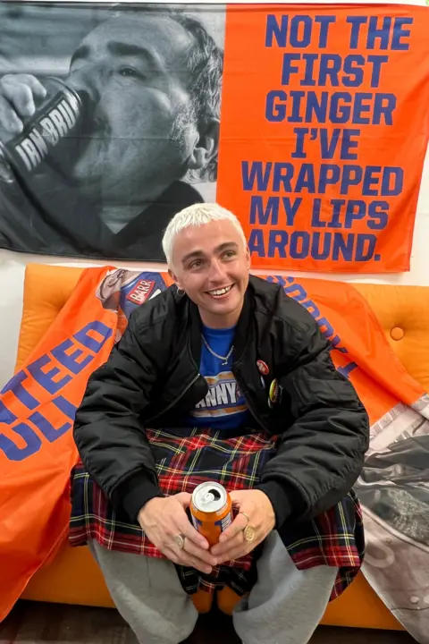 Trackie McLeod Trackie sits on a couch wearing a kilt and drinking a can of Irn-bru. Behind him is an Irn-bru flag that says "first ginger I've wrapped my lips around."