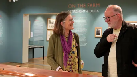 June Essex Sophie Mason, smiling and talking to Joe Pearson, in an exhibition. She is side on looking at him. Joe is wearing a white top, black jacket and has glasses on with short grey hair and a beard. Sophie is waring a dress, purple scarf, jacket and has shoulder length hair and is also wearing glasses.