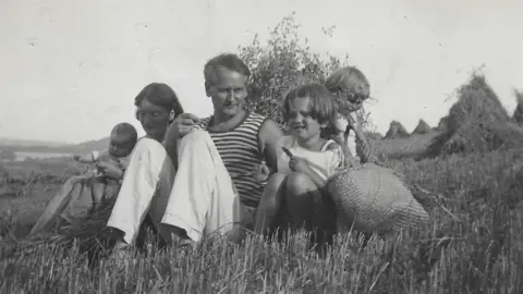 Family collection A black and white photo of Noel Carrington, with a woman and three children in a field. They are all sitting down with hay around them and a child holding a wicker bag. 