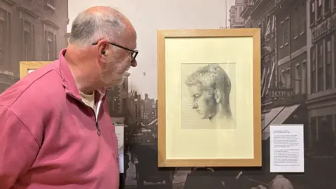 Victoria Partridge Joe Pearson, standing by a painting of Noel Carrington, which shows the side of a man's face, in black and white, placed on a picture of an old High Street. Joe is wearing a pink jumper, and has short grey hair and is wearing glasses. He is side on. 