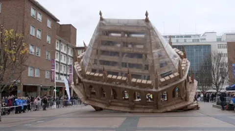 An art installation made of cardboard representing Coventry Cathedral. It is 15ft high and in a public square, surrounded by shops and members of the public. It is lying partially on its side. Crowds behind a barriers surround it.