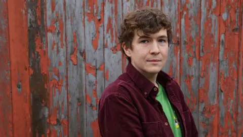 Colin Mearns/The Herald Novelist Callum McSorley, a brown-haired man in a purple cord shirt and green T-shirt looks along his shoulder, standing side-on.