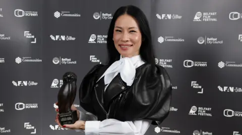 PRESS EYE Lucy Liu smiles while posing with her award at the Belfast Film Festival. She has long, straight, black hair and is wearing a black leather frock coat with puffed sleeves over a white blouse tied with a large bow at her neck. She is holding an award shaped like an owl. 