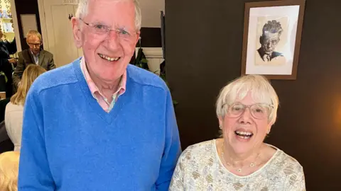 Angela O'Connor Two people standing indoors against a dark wall with a framed monochrome portrait hanging behind them. The person on the left is wearing a bright blue sweater over a collared shirt, while the person on the right is wearing a light patterned top.