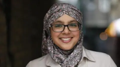 Rehan Jamil A woman in a floral headscarf, white coat, black glasses and pearl-like necklace smiles at the camera. The background behind her is blurred.